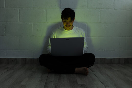 Man sitting on the floor of his home using a laptop at night, illuminated by the screen in a dark environment, relaxed and entertained while using technology from home