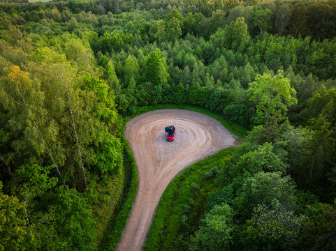 Aerial view of a red car with a roof tent parked in a circular dirt clearing surrounded by a dense green forest in Gavieze parish, South Kurzeme Municip, Latvia.