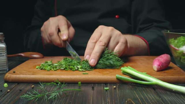 Chef prepares fresh herbs by chopping dill and green onions on a wooden cutting board with a pink radish and salad bowl in the background
