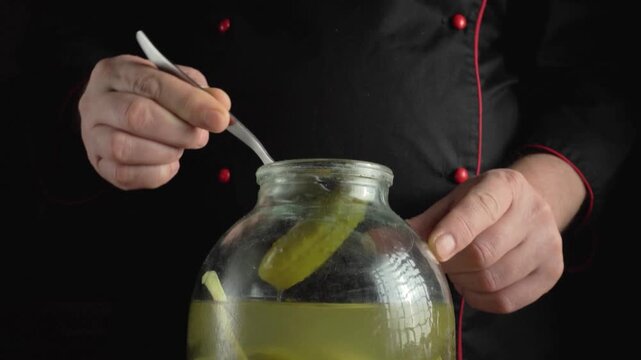 Chef in black uniform uses fork to lift pickle from jar, showcasing the process of preparing ingredients for a dish in a kitchen setting
