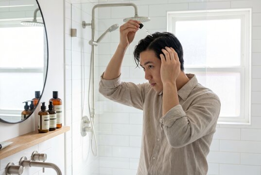 Young asian man applying serum to scalp in modern white bathroom. Routine haircare and grooming for healthy hair growth. Wellness lifestyle concept for skin and body cosmetic treatment.