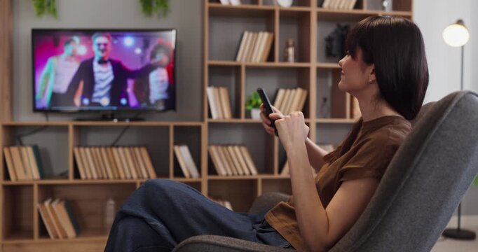 Woman watching television with remote on sofa. Cozy interior with shelving and TV, music show on screen, evoking leisure and weekend comfort. Home streaming relaxation and media entertainment.