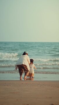 Back view of an Asian mother holding her toddler daughter's hand while standing on the sandy beach, looking at sea waves during sunset. Family bonding, parenting lifestyle, summer travel vacation.