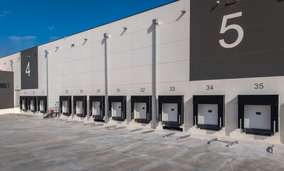 Naklejka premium Brand new large-scale logistics warehouse exterior showing numbered loading bays, modern concrete facade with bold signage, representing supply chain and e-commerce fulfillment infrastructure
