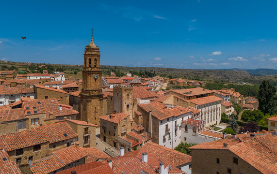 Elevated view of La Iglesuela del Cid in Maestrazgo, Teruel, Spain, featuring a baroque clock tower rising above terracotta-roofed stone buildings on an arid plateau under blue sky