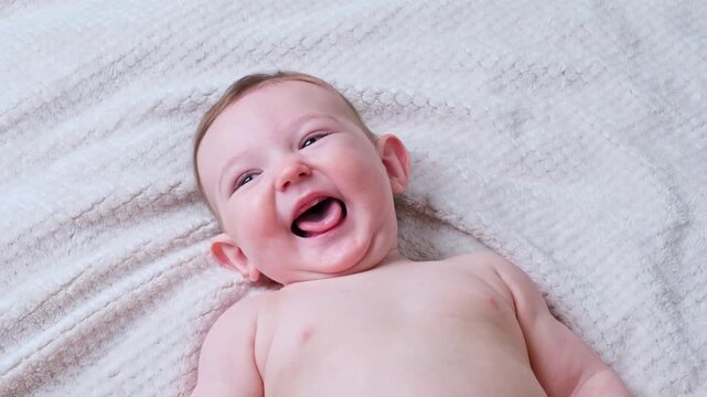 Laughing baby lying on soft white blanket. Joyful infant child showing toothless smile and playing. Excited toddler discovery and development concept. Innocent childhood emotion.