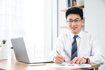 Fototapeta premium Smiling businessman taking notes with charts and laptop in modern office