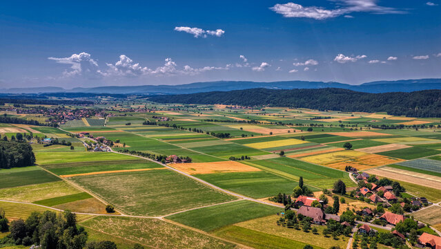 Panoramische Luftaufnahme einer l&auml;ndlichen Agrarlandschaft mit bunten Feldern, Bauernh&ouml;fen, D&ouml;rfern und Waldgebieten unter blauem Himmel mit Sonnenstrahlen