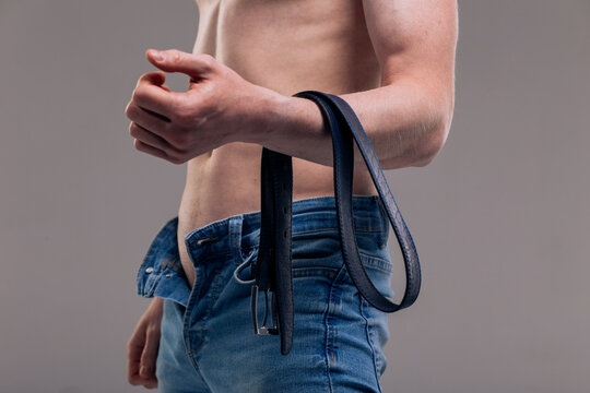 A muscular man in blue classic jeans with a bare torso is holding a belt. Concept: domination and play in a married couple, domestic violence, getting ready for work in the morning