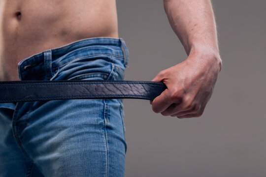 A muscular man in blue classic jeans with a bare torso is holding a belt. Concept: domination and play in a married couple, domestic violence, getting ready for work in the morning