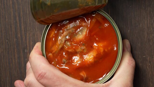 Close up of male hands opening a canned fish in tomato sauce pull ring lid over a dark wooden table. Conceptual backdrop for grocery retail, quick meals, emergency food supplies or cooking recipes