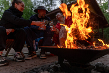 Three friends sit near glowing fire pit, holding drinks and talking beside rustic cabin in wooded setting, with warm flames lighting faces and courtyard © Egor Lint