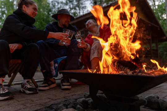 Three friends sit near glowing fire pit, holding drinks and talking beside rustic cabin in wooded setting, with warm flames lighting faces and courtyard
