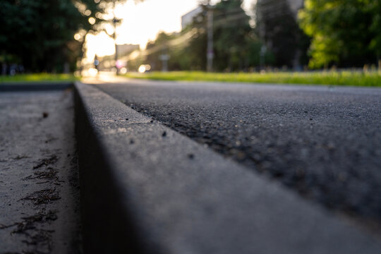 Low angle selective focus shot of a concrete curb stone and asphalt pathway with heavily blurred city street lights and trees at sunset.