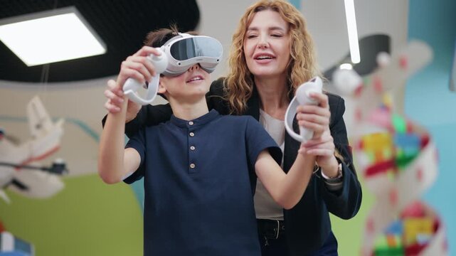 Tracking shot of boy wearing vr headset and holding controllers with woman guiding in colorful classroom playroom indoors.