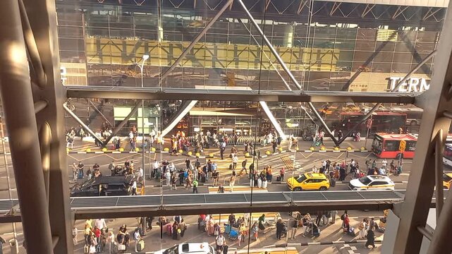 High angle shot through a big window of a modern airport terminal entrance with many travelers with suitcases on a crosswalk, yellow cabs, urban buses, and cars on the street outside the facility