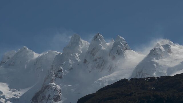 Close-up view of sharp, snow-covered mountain peaks, featuring dramatic windblown snow clouds swirling around the summits against a clear blue sky.