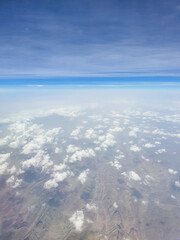 Fototapeta premium High-altitude aerial view of rugged mountain ridges partially covered by scattered clouds under a layered blue sky. View from a plane window