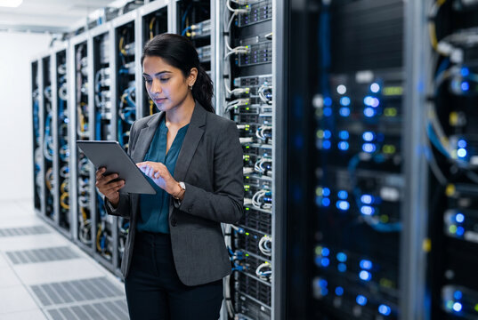 Female IT specialist using a digital tablet while working in a modern data center server room with rows of network equipment