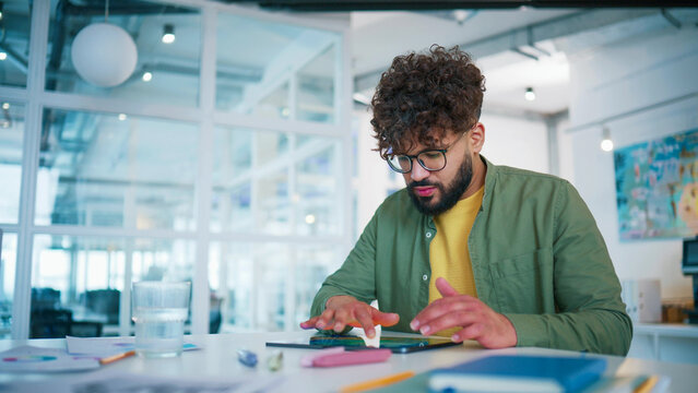 Focused male working on tablet at desk. Bearded man wearing glasses reviewing digital report. Touching screen and checking statistics. Glass of water nearby. Concentrating on financial analysis.