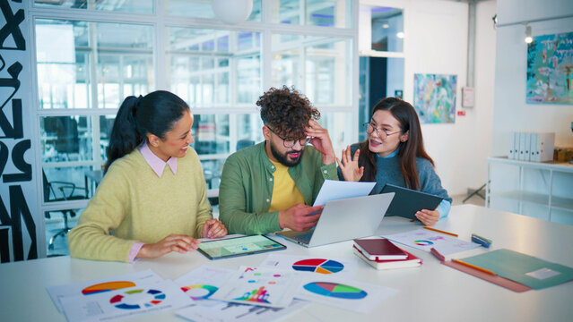 Business team sitting at table with financial charts. Group colleagues discussing project. Male reviewing printed documents. Female holding tablet and presenting ideas inside glass office meeting room