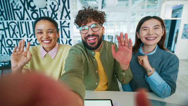 Cheerful office team taking selfie at workplace. Group colleagues extending arm and holding camera. Business people waving hands and smiling. Friendly workers greeting viewer in creative studio.