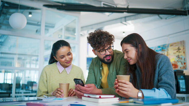 College friends gathering at desk with coffee cups. Group students watching funny video on smartphone. Male holding mobile device and sharing content. Classmates relaxing together in campus study area