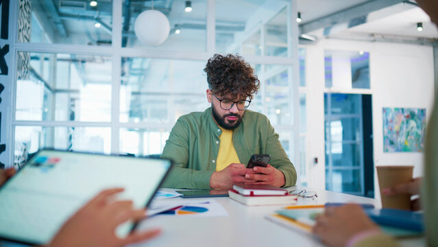 Focused male employee sitting at desk with notebook and tablet. Young man browsing content on smartphone. Worker holding mobile device and reading messages. Business professional working alone.