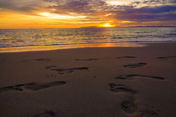 Obraz premium LENS FLARE: Numerous footprints trace paths along wide sandy shoreline under colorful sunset. Vibrant setting sun breaks through clouds and spreads a warm light across ocean tides on an island beach.