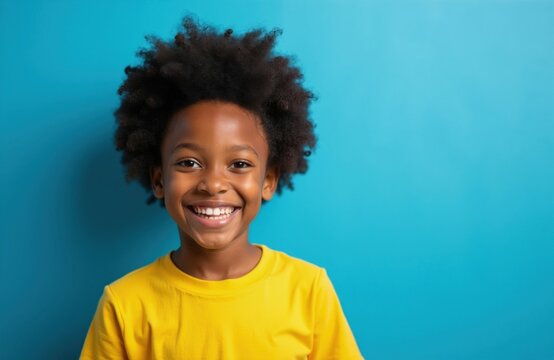 Young Black child with afro hair wears a bright yellow shirt. The kid smiles happily against a solid blue backdrop. This cheerful portrait conveys youthful innocence and vibrant energy.