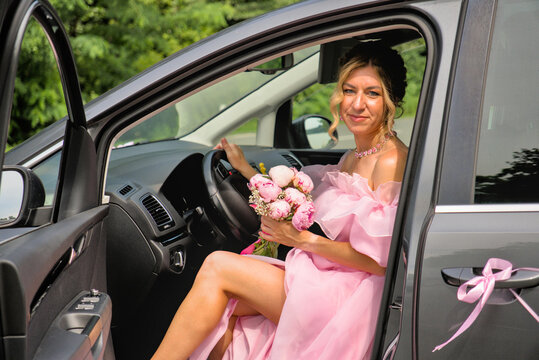 Elegant young woman in a fluffy pink evening gown sitting at the steering wheel of a wedding car.