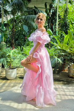 Fashion portrait of a beautiful woman in a voluminous pink gown standing full length among tropical plants in a greenhouse.