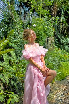 Dreamy young woman in a pink ball gown posing in a botanical garden among green palms, looking up at the sunlight.