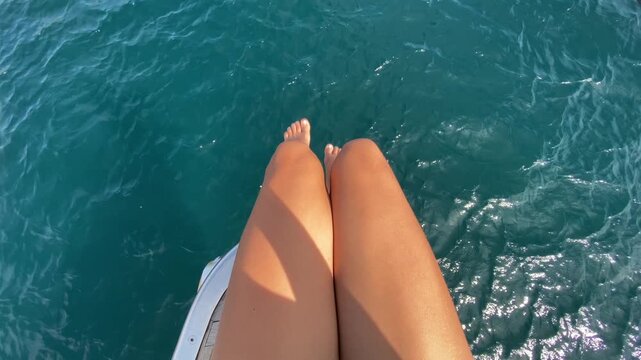Top-down view of a woman sitting on the bow of a yacht, gently swinging her legs above the crystal-clear turquoise waters of the Sea of Cortez. The boat glides smoothly across the calm sea.