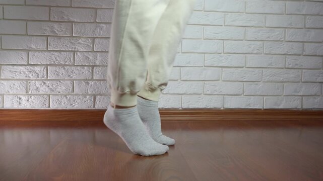 Woman stretching her feet and calves during home workout. Low angle view of woman's legs in sweatpants and socks performing calf raises and stretching her feet on a wooden floor, improving flexibility