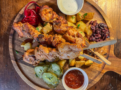 Wooden serving board with grilled meat skewers, roasted potatoes, pickled cucumbers, red onions, olives, and dipping sauces, creating a hearty rustic meal presentation
