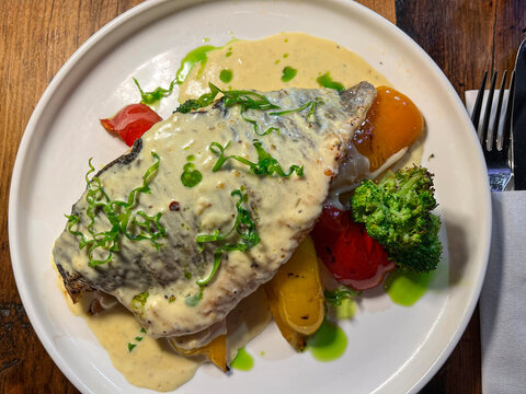 White plate featuring grilled Branzino sea bass fish fillet topped with creamy herb sauce, accompanied by roasted vegetables.