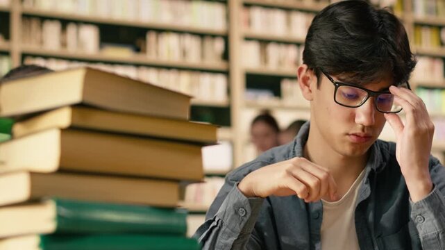 Exhausted asian student leans on one hand while reading open book in library, framed by stacks and green lamps during demanding study session at wooden desk.