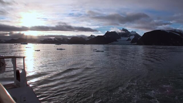 Exploring the sublime Arctic expanse of Svalbard, Norway. A vessel carves a path through shimmering waters, illuminated by the summer sun. Icebergs float near towering, glacier-etched peaks.
