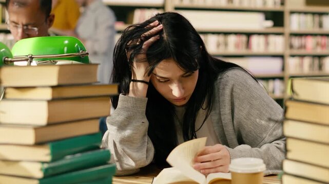 Exhausted young woman reading open book at university library desk, framed by stacks of books and coffee during long exam study session under pressure, Academic stress, with coursework and assignment.