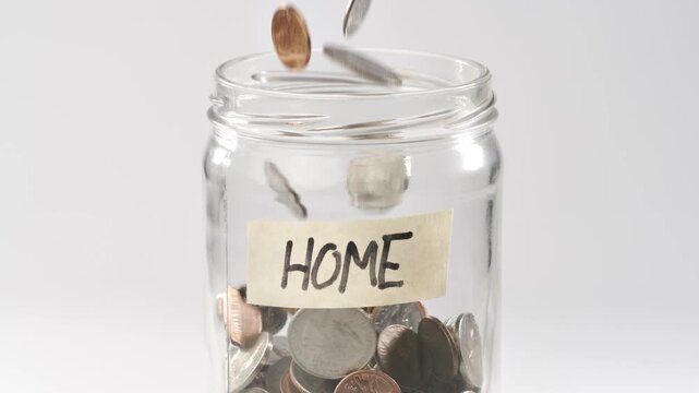 Close-up shot of coins falling in slow motion into a coin jar labeled 'Home'.