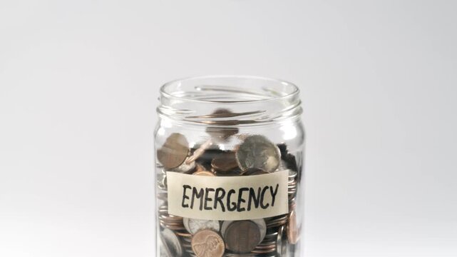 Close-up shot of coins falling into an 'Emergency Fund' coin jar in slow motion.