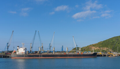 Naklejka premium Large commercial bulk carrier ship docked at an industrial sea port with loading cranes and green hills. Represents global trade and logistics