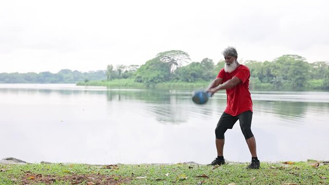 Mature Indian man with a beard working out with a kettlebell in a lakeside park In Singapore.