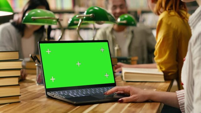 Green screen mockup on laptop inside university library desk, study space surrounded by blurred students and green lamps, Editable screen on device with touchpad, with group study and assignment.
