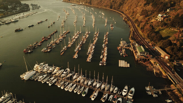 Aerial view of the marina with rows of sailboats and yachts moored on the River Dart, featuring a railway line along the wooded bank in Kingswear, England, United Kingdom.