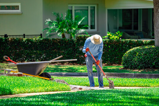 Professional landscaper using a shovel to edge a walkway on a lush green residential lawn next to a wheelbarrow