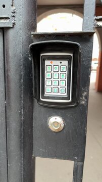 Analog intercom keypad on iron gate, weathered black frame, green backlit numeric buttons, round scanning key below keypad, brick