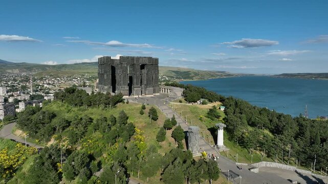 Drone shot capturing the massive bronze and stone pillars of the Chronicle of Georgia monument complex, situated on a green hill overlooking the vast Tbilisi Sea reservoir under a clear blue sky