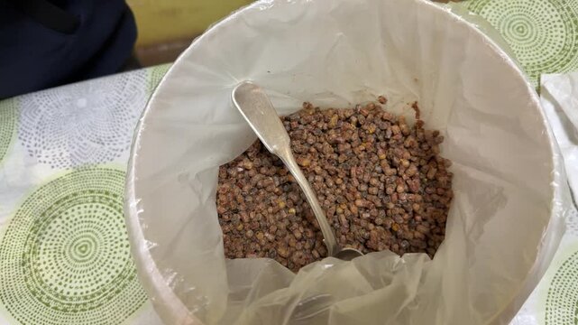 Close-up bee pollen in bucket lined with plastic bag and metal spoon, golden brown granules and clusters, earthy texture